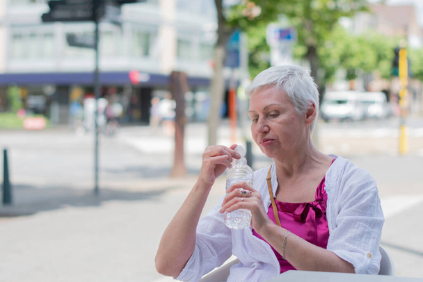 60歳の白人女性ボブがスタイリッシュな夏服で切り取り街のカフェテーブルでペットボトルから水を飲む高品質の写真 - 写真・画像