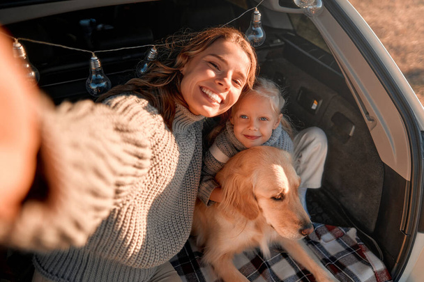 Μια νεαρή γυναίκα μητέρα με την χαριτωμένη κόρη της και το σκύλο της να κάθεται σε μια ζεστή κουβέρτα στο ανοιχτό πορτ-μπαγκάζ ενός αυτοκινήτου και να βγάζει μια σέλφι στο τηλέφωνο. Οικογένεια ταξιδεύει με ένα κατοικίδιο ζώο. - Φωτογραφία, εικόνα