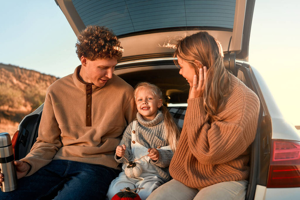 一緒に楽しい週末に毛布の上に車の後ろに座っている彼らの最愛の小さな娘とピクニック上の若い幸せな家族. - 写真・画像