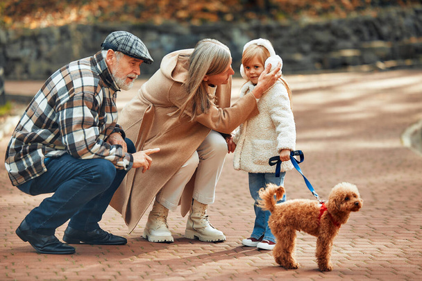 Reifes Paar, das mit Kind und Hund im Park spazieren geht. Großeltern gehen im Herbst mit Enkelin und Hund im Park spazieren. - Foto, Bild