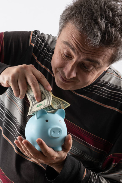 Ein Mann mittleren Alters mit grauen Haaren in lässiger Kleidung versucht, 1-Dollar-Scheine in ein Sparschwein zu legen - Foto, Bild