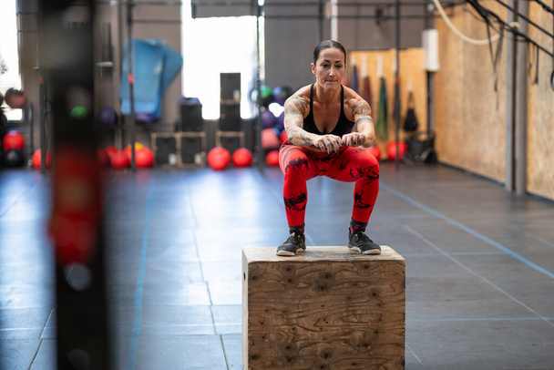 Strong tattooed woman jumping on a box in a cross training center - Photo, Image