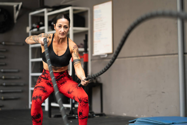 Strong mature woman working out with battle ropes in a cross training gym - Photo, Image