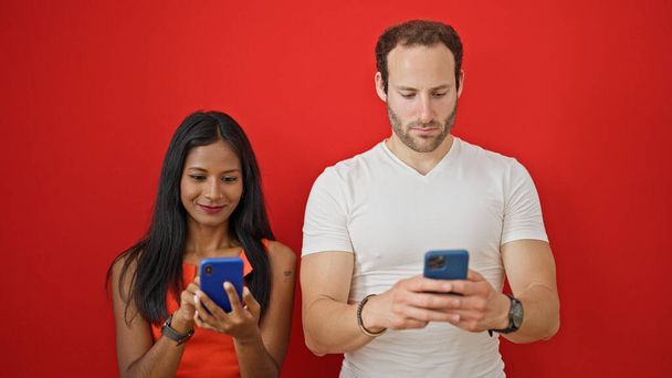 Όμορφο ζευγάρι που χρησιμοποιεί smartphones χαμογελώντας πάνω από απομονωμένο κόκκινο φόντο - Φωτογραφία, εικόνα