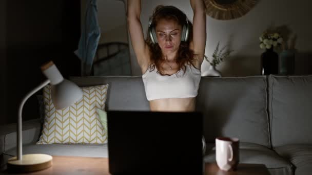 Una giovane donna in cuffia si allunga mentre lavora fino a tardi su un computer portatile nel suo soggiorno - Filmati, video
