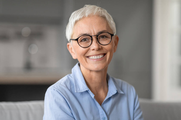 Portrait of happy caucasian mature woman posing on sofa at home interior, looking and smiling at camera. Cheerful woman in stylish eyewear expressing positivity and confidence - Photo, Image