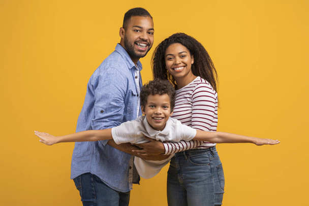 Οικογενειακό δέσιμο. Χαρούμενος μαύρος άντρας και γυναίκα να κρατούν το γιο τους στα χέρια, χαμογελαστό αγόρι να προσποιείται ότι πετάει, να απλώνει τα χέρια, να μιμείται το αεροπλάνο, να διασκεδάζει με τους γονείς του σε κίτρινο φόντο στούντιο - Φωτογραφία, εικόνα