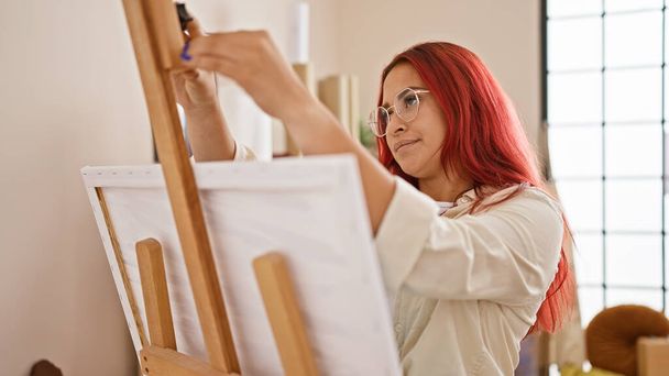 Lebendige junge rothaarige Künstlerin voller Kreativität, die sich freudig auf das Malen vorbereitet, inmitten ihres Ateliers steht, bereit, Leinwand auf Staffelei zu stellen und ihr Meisterwerk zu zeichnen - Foto, Bild