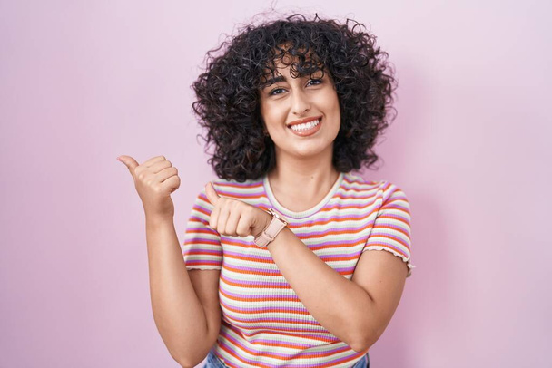 Young middle east woman standing over pink background pointing to the back behind with hand and thumbs up, smiling confident  - Photo, Image