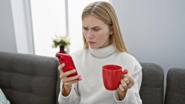 Une femme blonde surprise tenant une tasse rouge et un smartphone réagit au contenu de son appartement. - Séquence, vidéo