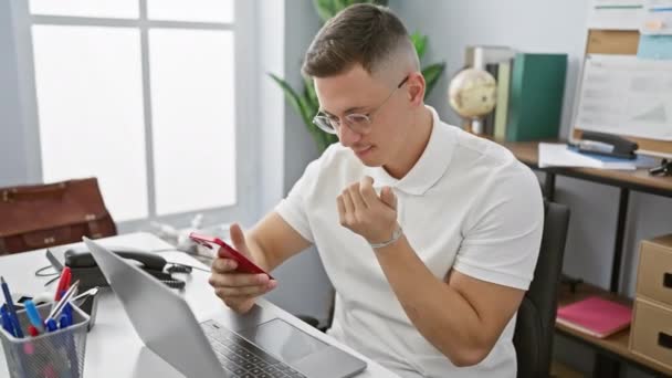 Un joven hispano trabaja en su portátil en una oficina moderna, analizando documentos mientras sostiene su teléfono inteligente. - Imágenes, Vídeo