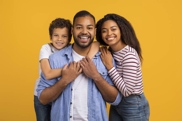 Feliz família negra de três abraços enquanto posam juntos em estúdio, pais americanos africanos alegres e pequeno filho compartilhando abraço afetuoso, sorrindo para a câmera, de pé contra o fundo amarelo - Foto, Imagem
