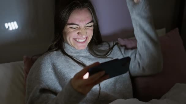 Usmívající se mladá hispánka se těší pomocí svého smartphonu ve své ložnici v noci. - Záběry, video