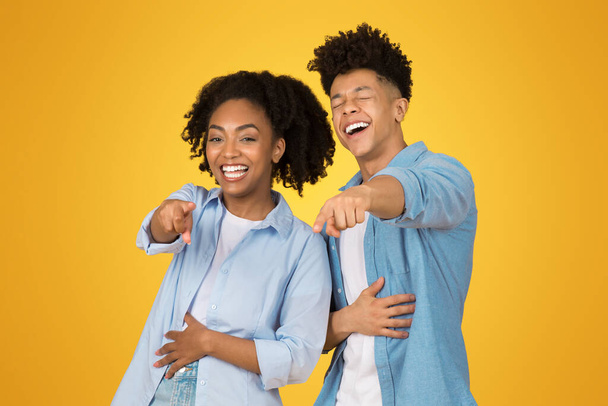 Herzhaft lachend stehen eine junge afrikanisch-amerikanische Frau und ein junger Mann dicht beieinander und zeigen in die Kamera, als teilten sie einen Witz, vor einem fröhlich gelben Hintergrund. - Foto, Bild