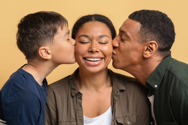 Boldog Anyák Napját! Szerető fekete férfi apa és tinédzser fiú csókolózik boldog fiatal nő, közelkép, elszigetelt színes háttérrel. Aranyos család ünnepli a nemzetközi nőnap - Fotó, kép