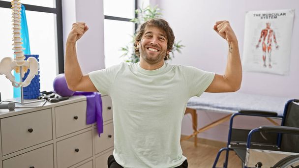 Un joven alegre flexiona los músculos alegremente en la sala de terapia de una clínica de rehabilitación bien equipada. - Foto, Imagen