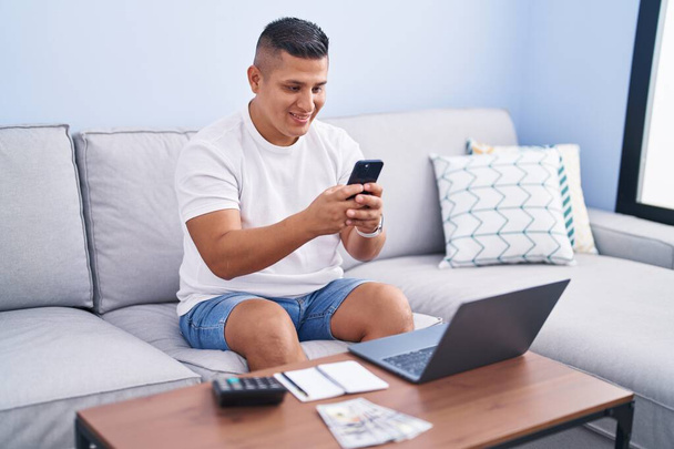 Giovane uomo latino utilizzando smartphone e laptop a casa - Foto, immagini