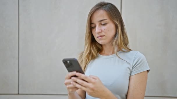 Vrolijke jonge blonde vrouw gelukkig aanraken van haar smartphone, bezig met een digitaal gesprek op een zonnige straat - Video