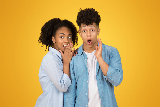Une jeune femme afro-américaine murmure dans sa main avec une expression espiègle tandis qu'un jeune homme à côté d'elle réagit avec une main sur sa joue, tous deux sur un fond jaune - Photo, image
