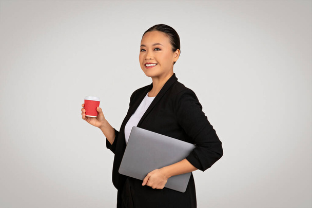 Profissional mulher de negócios asiática segurando um laptop sob seu braço e uma xícara de café vermelho, pronto para um dia produtivo no trabalho com uma atitude positiva, isolado em fundo cinza, estúdio - Foto, Imagem