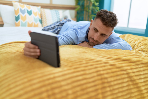Молодий іспаномовний чоловік, використовуючи тачпад, лежить на ліжку в спальні
 - Фото, зображення