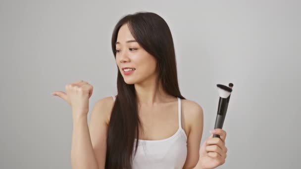 Neşeli genç Çinli kadın makyaj fırçalarını güvenle sergiliyor, başparmaklarını kaldırıyor ve saf beyaz izole bir arka planda neşeli, açık ağızlı bir gülümsemeyle kenarı işaret ediyor. - Video, Çekim