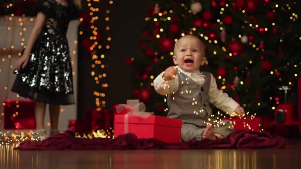 Egy aranyos fiú és a húga boldogok a szilveszteri ajándékokkal egy díszített karácsonyfa hátterében.. - Felvétel, videó