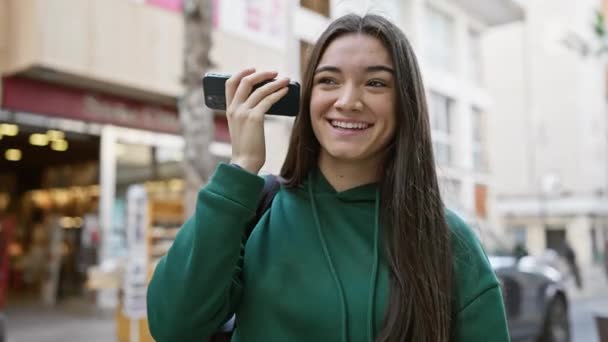 Een glimlachende jonge Spaanse vrouw die een telefoon tegen haar oor houdt in een stedelijke straat.. - Video
