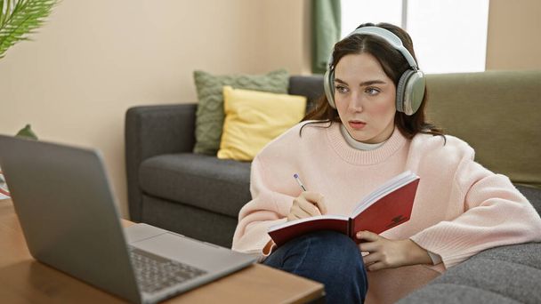 Kulaklıklı genç bir kadın rahat bir oturma odasında dizüstü bilgisayarla ders çalışıyor.. - Fotoğraf, Görsel
