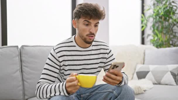 Homem árabe jovem grave duvidando sobre a mensagem de texto em casa, sentado dentro de casa, usando seu smartphone. relaxante com uma bebida de café expresso forte, perdido em pensamento profundo. - Filmagem, Vídeo