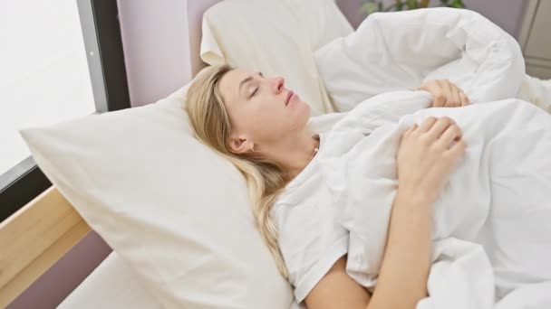 Una giovane donna bionda rilassata a letto con uno smartphone, che ritrae il tempo libero mattutino a casa - Filmati, video
