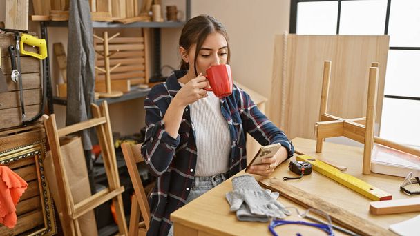 Mujer hispana disfruta del café mientras escribe en un taller de carpintería. - Foto, Imagen