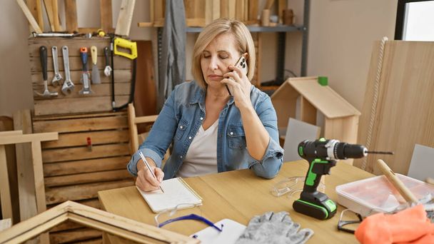 Una mujer de mediana edad multitarea en un taller de carpintería, hablando por teléfono mientras toma notas. - Foto, Imagen