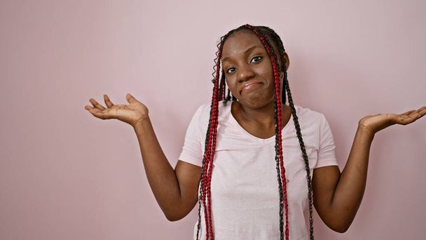 Ahnungslose afrikanisch-amerikanische Frau mit Zöpfen, die unsicher über einem rosa isolierten Hintergrund steht und Zweifel, Verwirrung und Gleichgültigkeit durch ihre Mimik ausdrückt - Foto, Bild