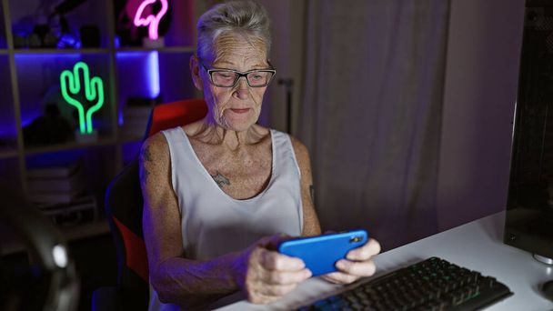 Gris cheveux, femme senior streamer apportant la chaleur, jouer à un jeu vidéo futuriste via smartphone dans la salle de jeux sombre à la maison - Photo, image