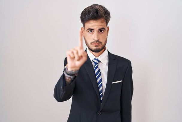 Mladý hispánec s tetováním v obleku a kravatou ukazující prstem nahoru a rozzlobeným výrazem, neprojevuje žádné gesto  - Fotografie, Obrázek