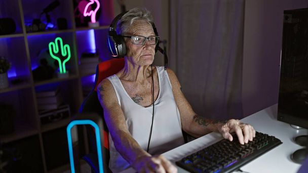 Serieux streamer femme âgée aux cheveux gris absorbé dans un jeu numérique nocturne à la maison, détendu mais concentré, assis avec casque et manette dans sa salle de jeu - Photo, image