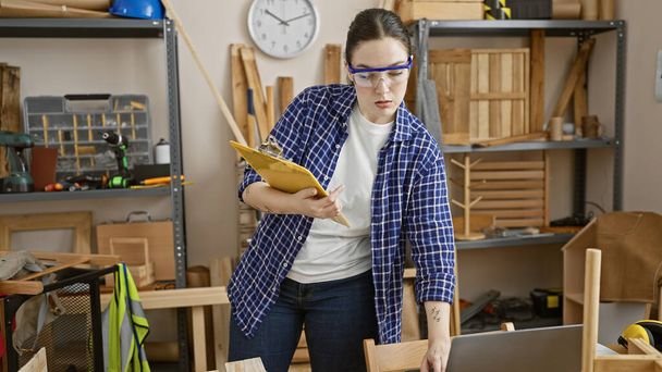 Mujer caucásica inspecciona portapapeles en un taller lleno de herramientas de carpintería y muebles. - Foto, Imagen
