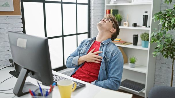 Overwerkte knappe jonge Latijns-Amerikaanse man werkt op kantoor business table, gekwetst door een plotselinge hartaanval, terwijl het nippen van koffie, stress en angst schijnbaar in zijn bril weerspiegelen - Foto, afbeelding