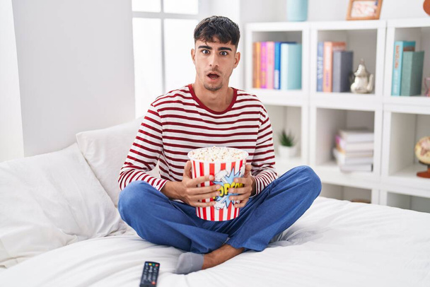 Молодий іспаномовний чоловік, що їсть попкорн, сидить на ліжку, дивлячись фільм в шоковому обличчі, виглядає скептично і саркастично, здивований відкритим ротом  - Фото, зображення