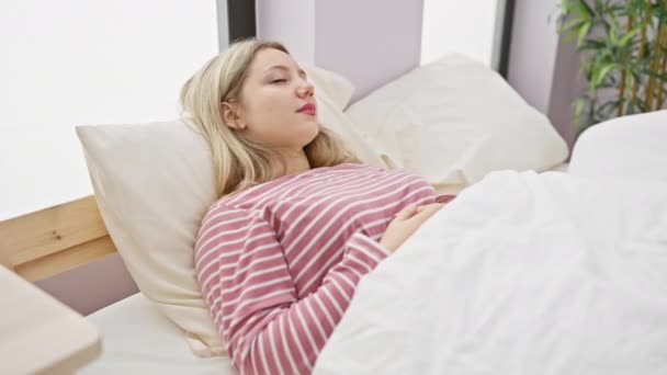 Een jonge blonde vrouw in roze gestreepte pyjama geniet van een ontspannen ochtend in een lichte slaapkamer, uitrekken en glimlachen met energie. - Video