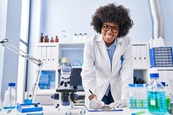 Femme scientifique afro-américaine souriante écriture confiante sur le document au laboratoire - Photo, image