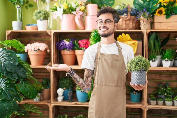 Young hispanic man with tattoos working at florist shop holding plant celebrating achievement with happy smile and winner expression with raised hand  - Photo, Image