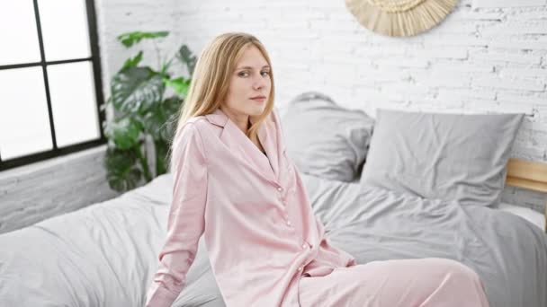 Een serene jonge vrouw in roze pyjama ontspannen in een lichte slaapkamer met een plant en bakstenen muur op de achtergrond. - Video