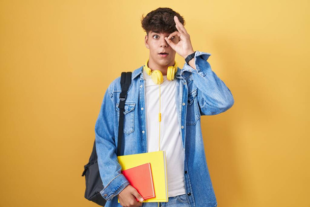 Hispanic teenager wearing student backpack and holding books doing ok gesture shocked with surprised face, eye looking through fingers. unbelieving expression.  - Фото, зображення