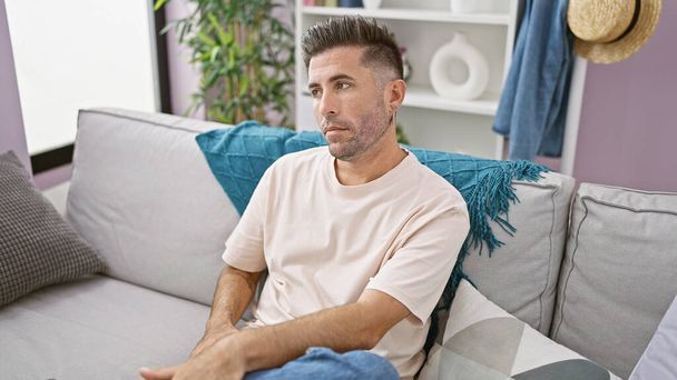 Handsome young hispanic man with a serious expression, sitting in comfort on a cozy sofa, relaxing and resting in his stylish indoor apartment living room at home - Photo, Image