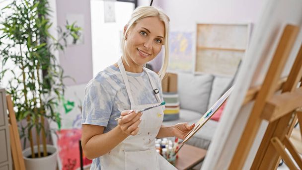 Jovem artista loira feliz, confiante e sorridente, desfrutando de sua lição de arte dentro de casa no estúdio de arte da universidade, enquanto desenha belas telas com pincéis - Foto, Imagem
