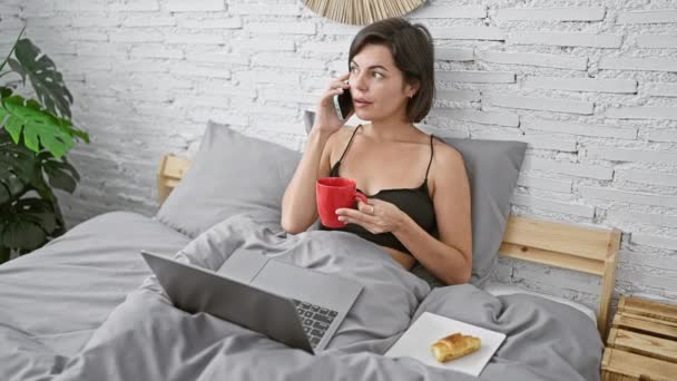 Strahlende junge hispanische Frau, die fröhlich am Telefon plaudert, während sie im Laptop surft, und den Morgenkaffee im hell erleuchteten Schlafzimmer genießt - Filmmaterial, Video