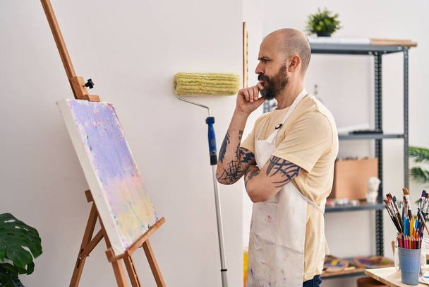 Jeune homme chauve artiste regardant dessiner avec une expression sérieuse au studio d'art - Photo, image