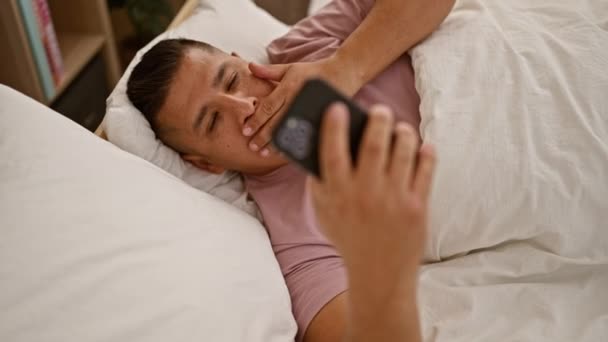 Knappe jonge Latijnse man, comfortabel liggend in zijn gezellige slaapkamer, het afronden van een ontspannen sms-sessie op zijn smartphone voordat je gaat slapen - Video